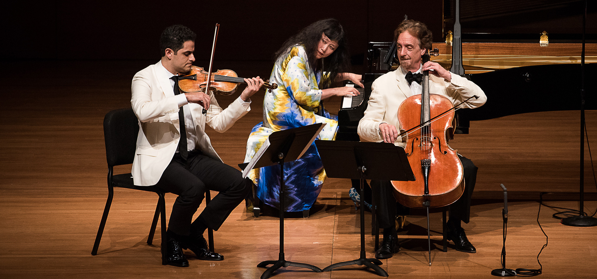 Three musicians performing seated together on stage; Wu Han on piano, David Finckel on cello, and Arnaud Saussman on violin.
