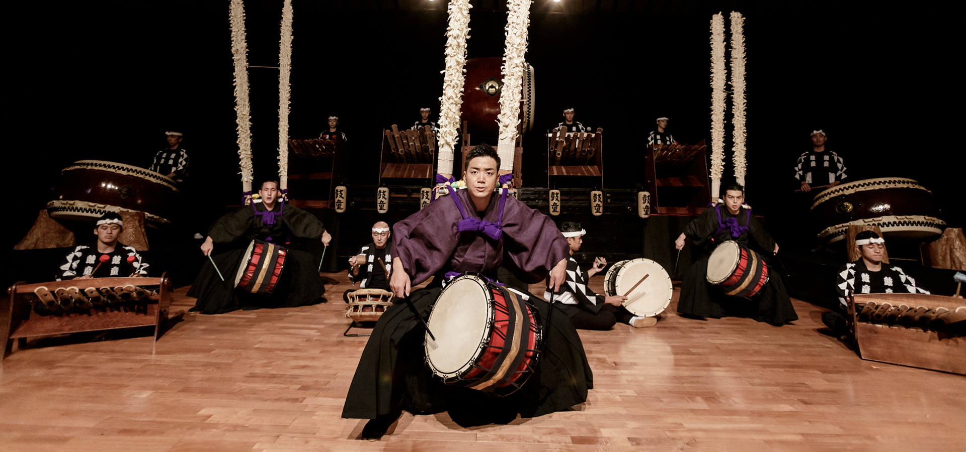 Members of the Kodo Troupe, wearing traditional Japanese attire, sit in front of numerous large drums on stage during an enthusiastic performance of Warabe.