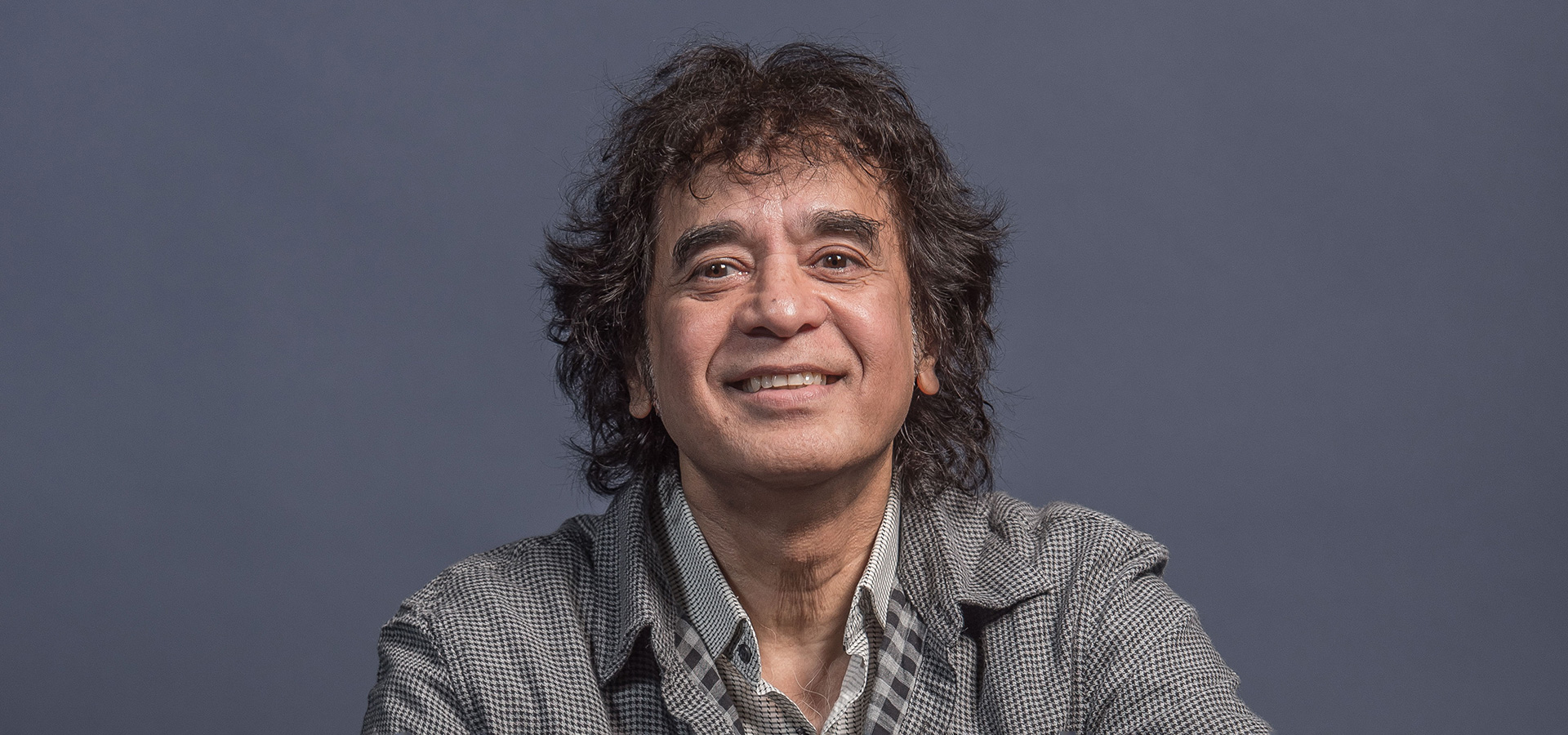 Zakir Hussain, a man with dark curly hair and bright brown eyes, smiles for the camera against a cool grey background.