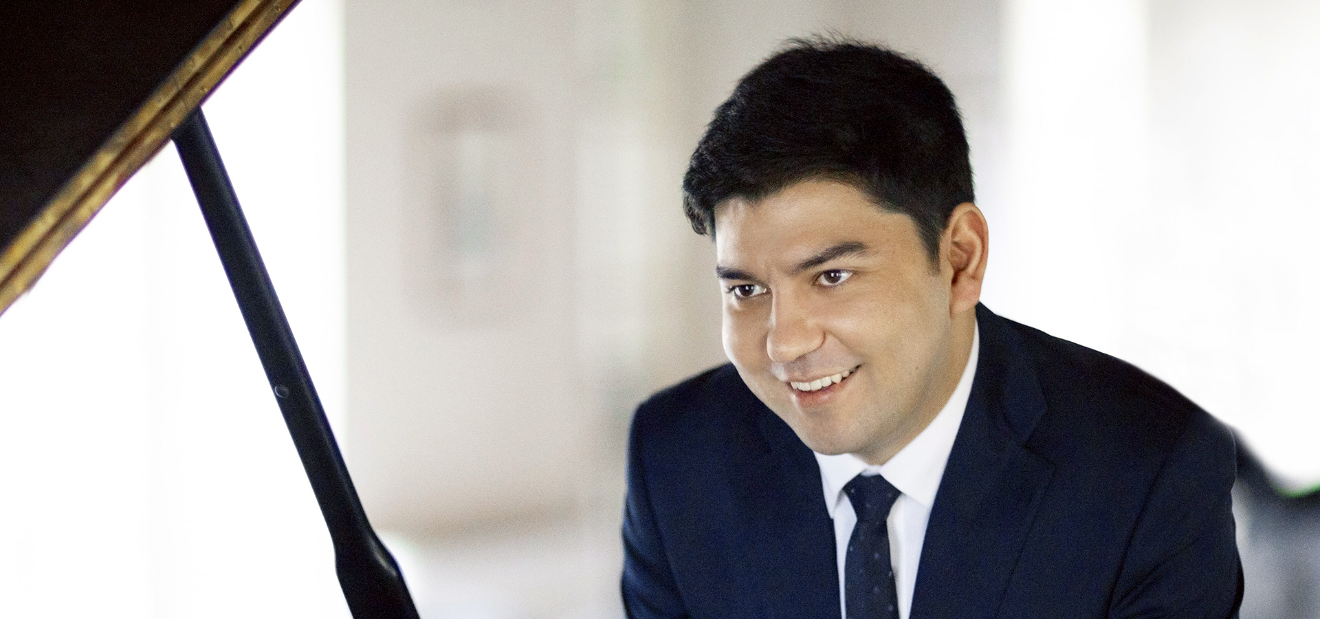 Behzod Abduraimov wears a navy suit and sits at an open grand piano, smiling.