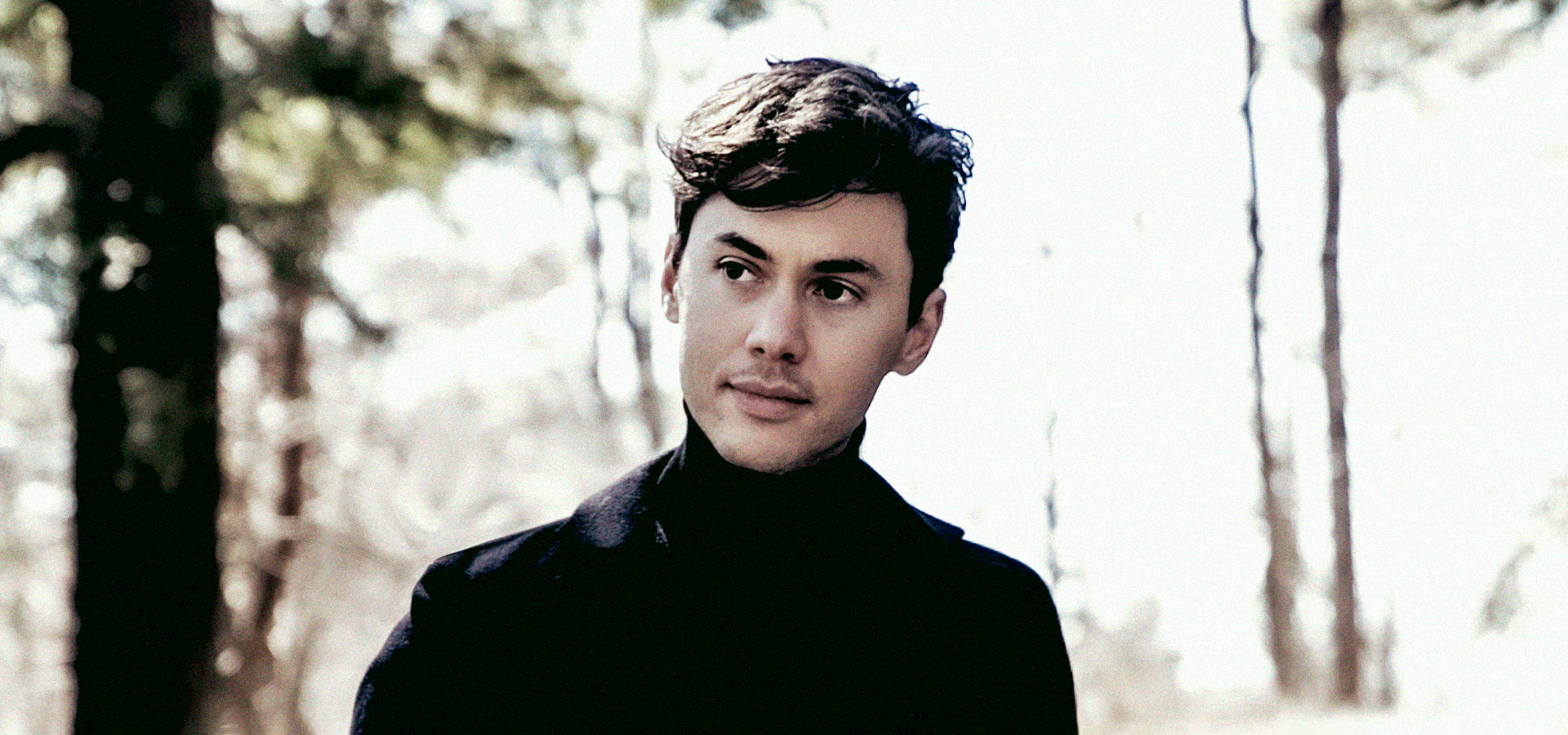 A portrait photo of violinist Benjamin Beilman, a young man wearing a black suit and turtleneck.