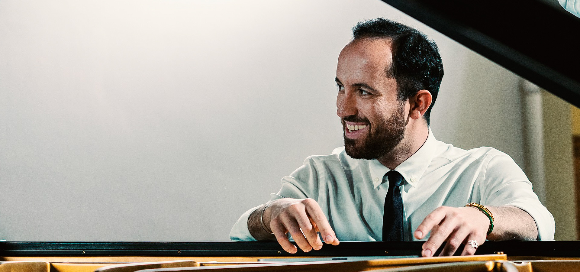 Igor Levit smiles and looks to his right as he sits in front of a piano in a white dress shirt and black tie.