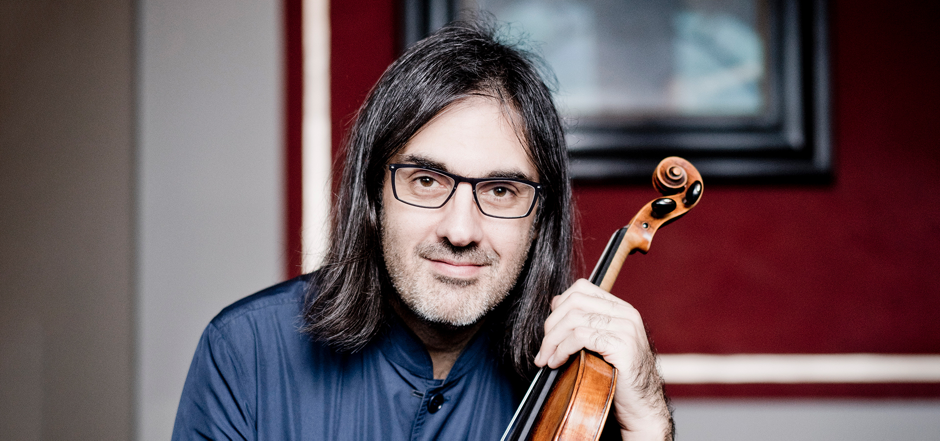 Leonidas Kavakos, a white man with dark shoulder-length hair and glasses, holds his violin and looks into the camera.