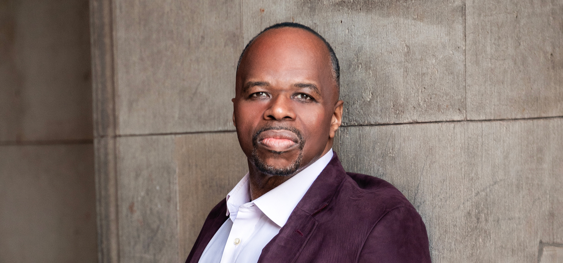 Lester Lynch, a Black man in a wine-colored jacket and pale pink shirt, rests against a light stone wall