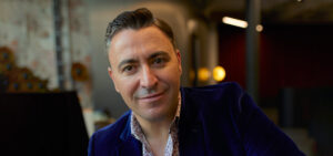 A close up photo of Maxim Venegrov, a middle-aged man wearing a blue velvet jacket and smiling at the camera.