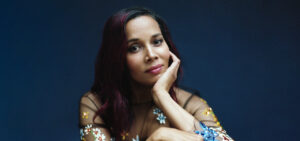 Rhiannon Giddens, a woman with long reddish-dark hair, sits looking at the camera in a transparent black top adorned with colored flowers.