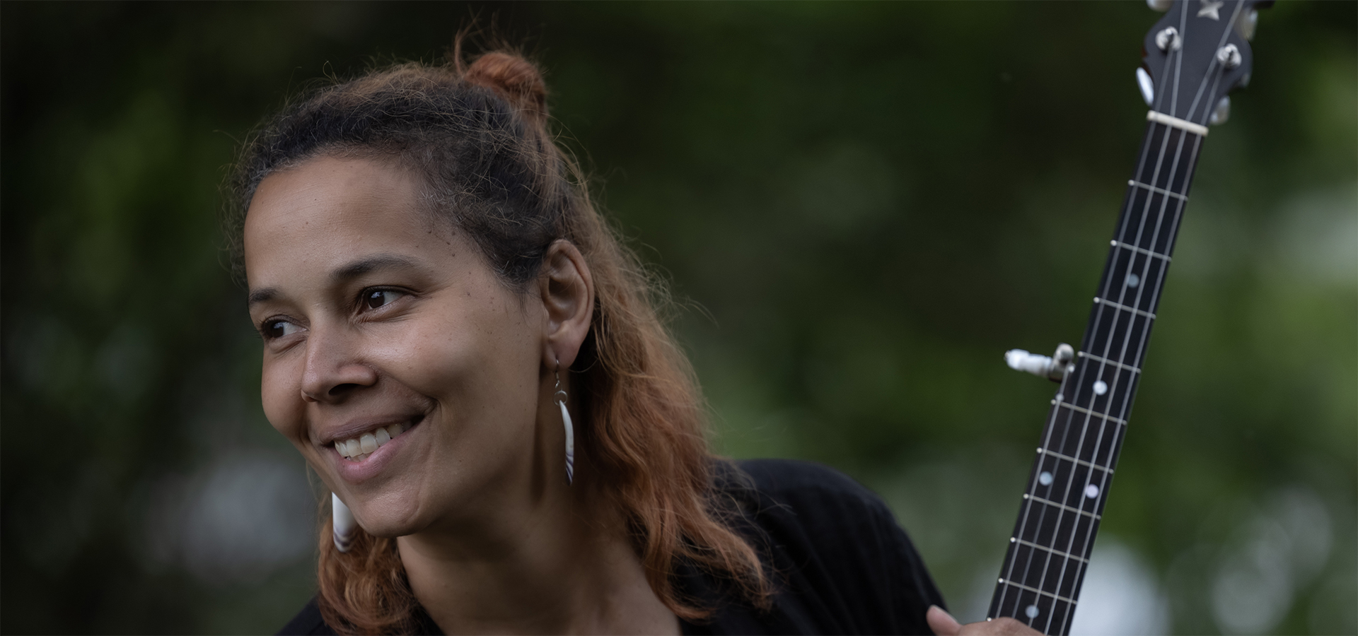 Rhiannon Giddens, a tan woman with wavy brown and orange hair half tied up into a bun, looks at something off to her side, while holding a banjo