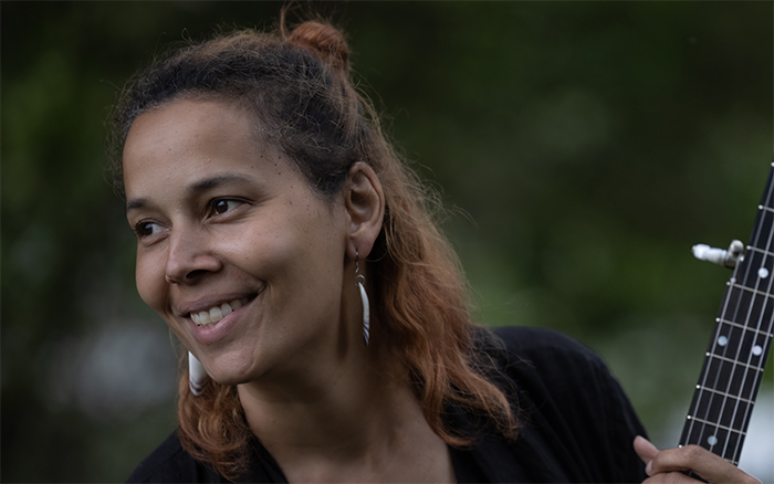 Rhiannon Giddens, a tan woman with wavy brown and orange hair half tied up into a bun, looks at something off to her side, while holding a banjo