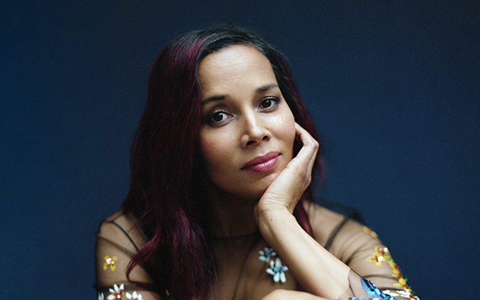 Rhiannon Giddens, a woman with long reddish-dark hair, sits looking at the camera in a transparent black top adorned with colored flowers.