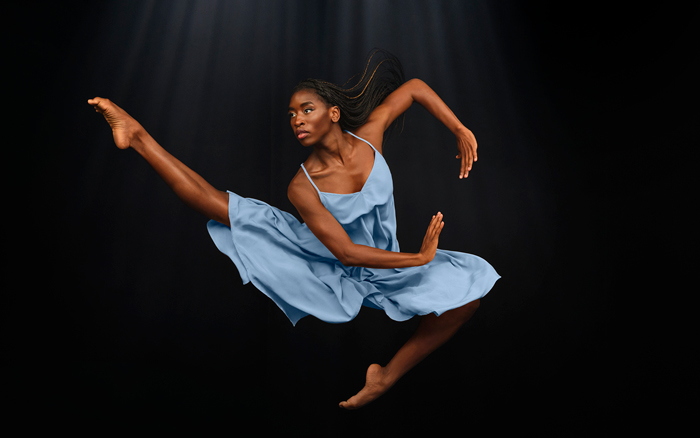 Photo of a Black female Alvin Ailey American Dance Theater dancer leaping in the air in a light blue dress