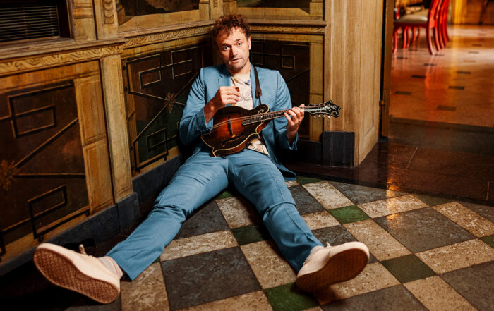 Chris Thile, a man with short brown hair wearing a light blue suit and white athletic shoes, slouches on the floor in an ornate room looking at the camera as he holds his mandolin.