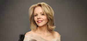 Renee Fleming, an older white woman with blond hair in an off-the-shoulder dress, smiles into the camera in front of a gray background.