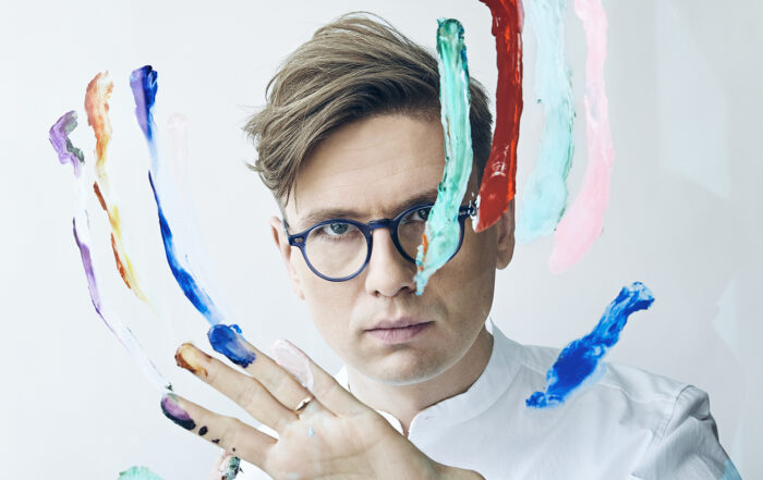 Vikingur Olafsson, an Icelandic man wearing blue rimmed glasses stares into the camera while smearing paint with his hands on a clear screen in front of him.