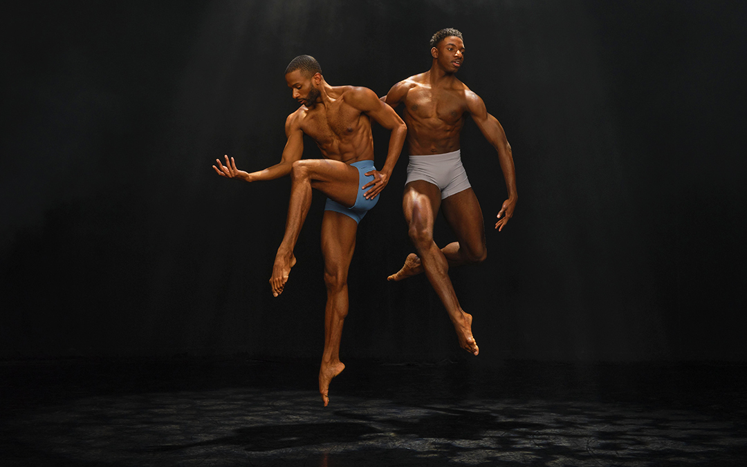 Two male dancers of Ailvin Ailey American Dance Theater wearing fitted shorts perform mid-air leaps against a dark background with overhead lighting highlighting their muscular physiques.