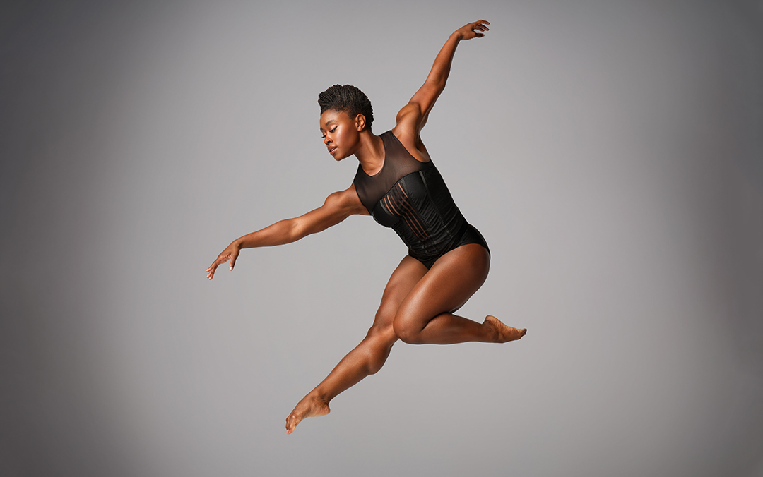 A solo female dancer of Ailvin Ailey American Dance Theater wearing a black sleeveless leotard performs a graceful mid-air leap against a gray background