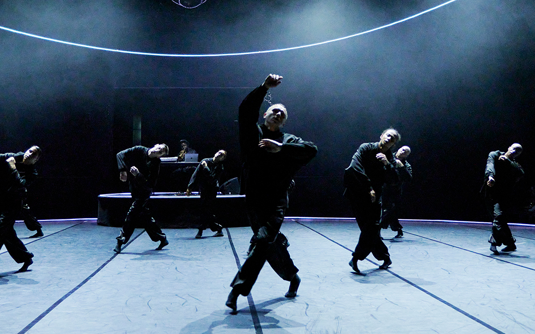 A group of dancers in B E N D in black outfits perform synchronized movements on a stage with a DJ in the background and curved lines of light above them.