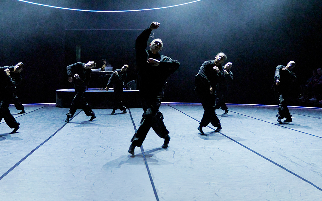 A group of dancers in B E N D in black outfits perform synchronized movements on a stage with a DJ in the background and curved lines of light above them.