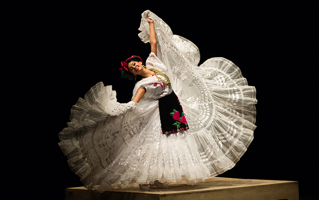 A solo woman poses in a traditional Mexican ornate flowing white dress, with her arms spead wide and the skirt in her hands, and smiles into the camera.