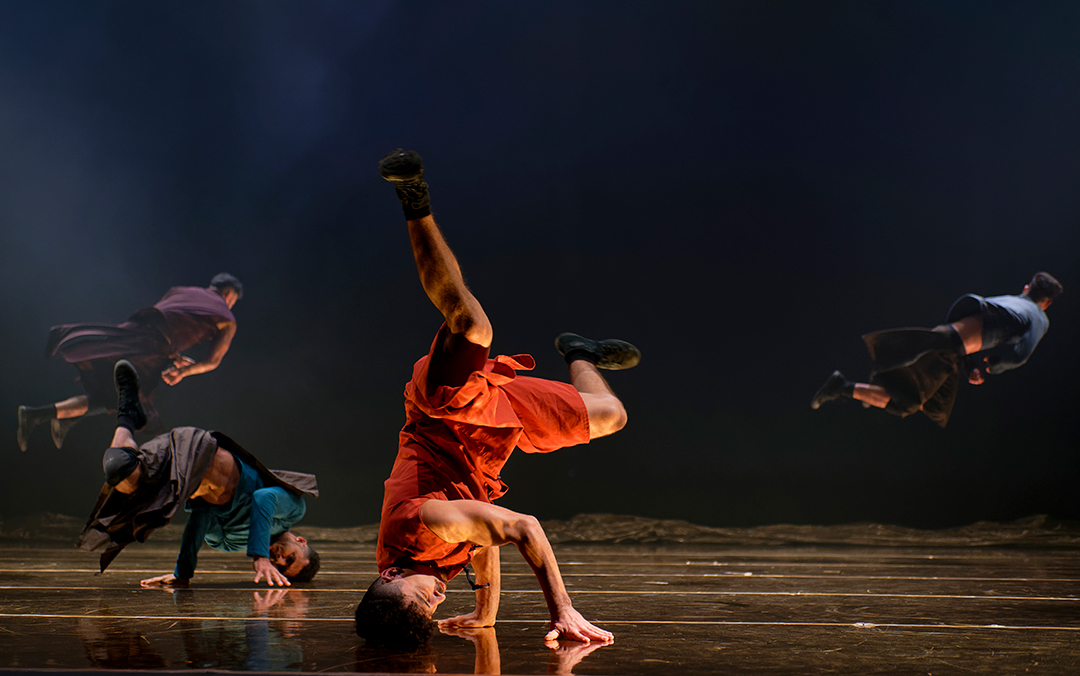 Several dancers of Compagnie Hervé Koubi performing on stage, several on their heads, displaying synchronized movements and colorful costumes.