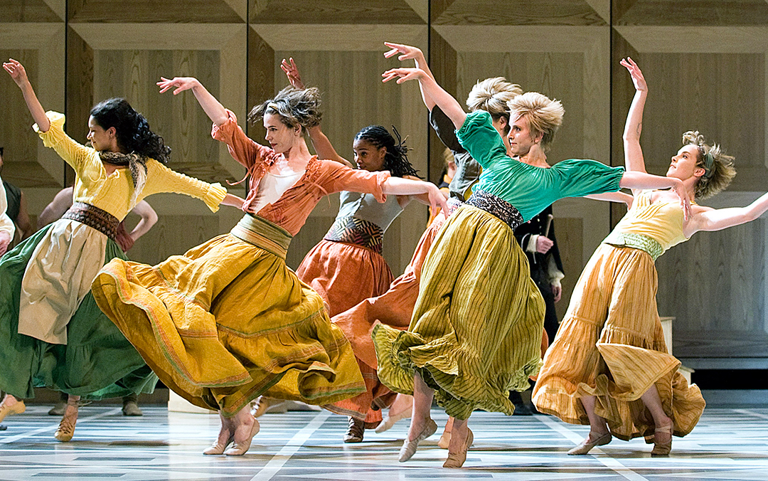 A group of Mark Morris Dance Group dancers in vibrant dresses performing on stage, displaying synchronized movements and lively expressions.