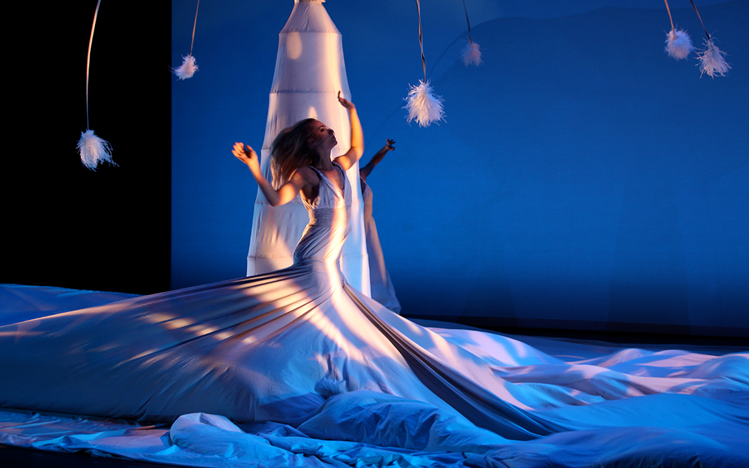 A solo female MOMIX dancer on stage in a wrapping, flowing white dress that covers most of the ground with her arms in the air and her head thrown back.
