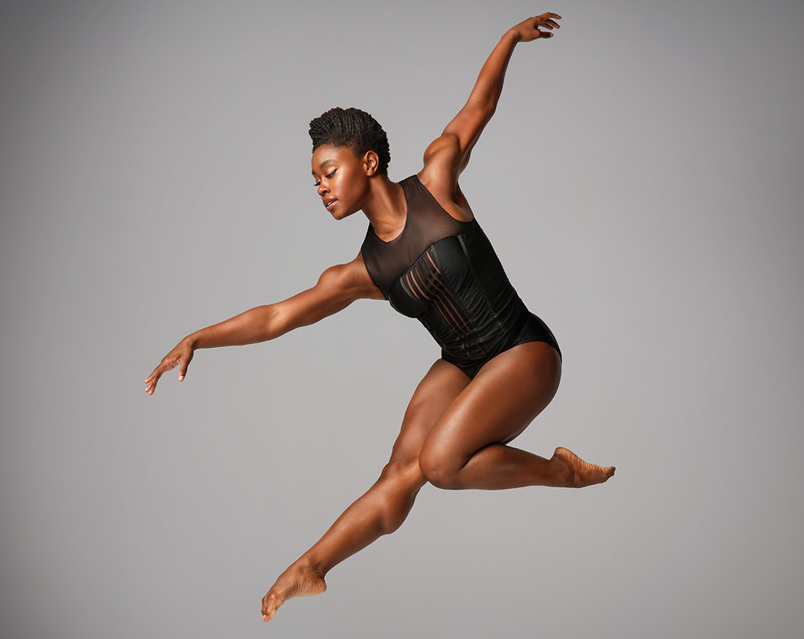 A solo female dancer of Ailvin Ailey American Dance Theater wearing a black sleeveless leotard performs a graceful mid-air leap against a gray background