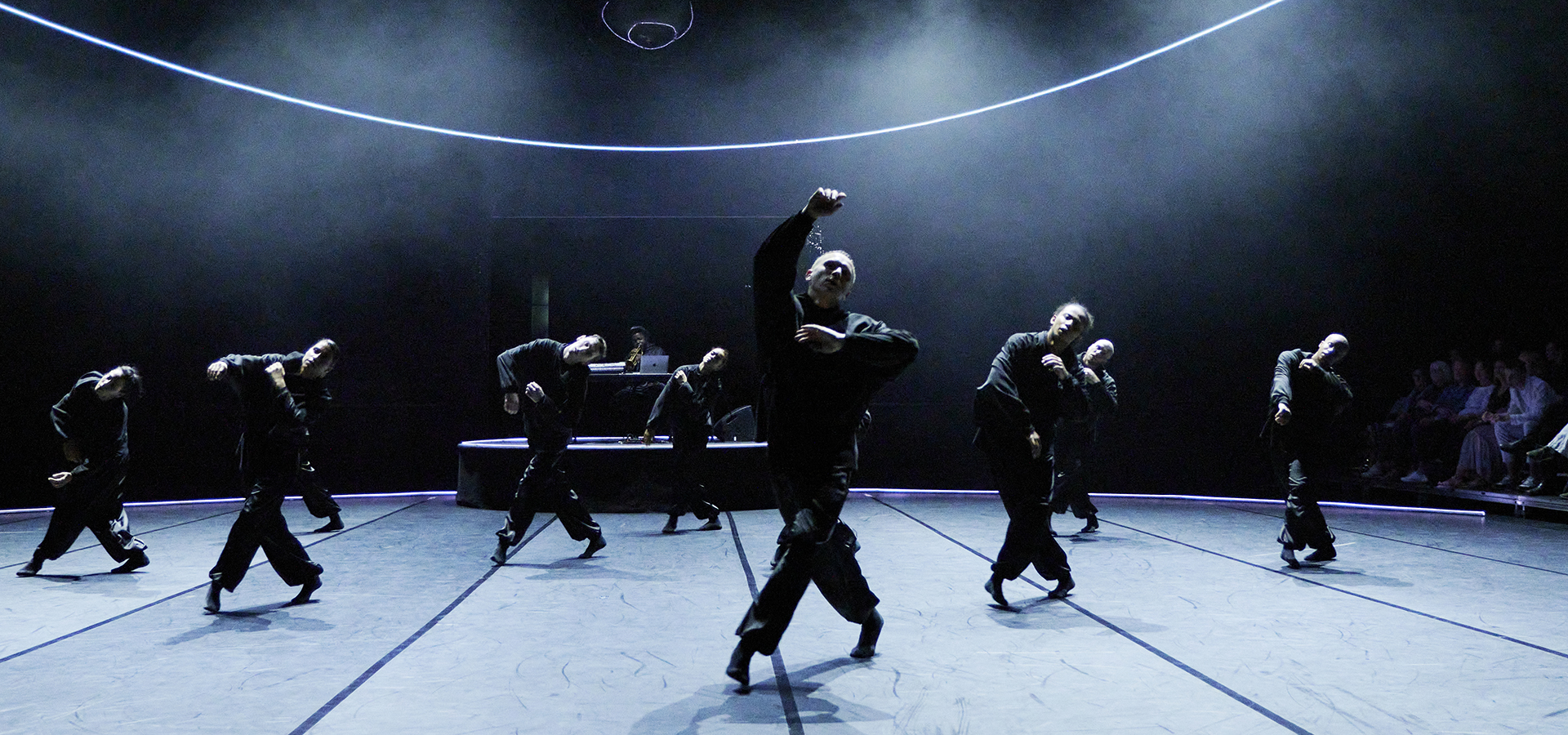 A group of dancers in B E N D in black outfits perform synchronized movements on a stage with a DJ in the background and curved lines of light above them.