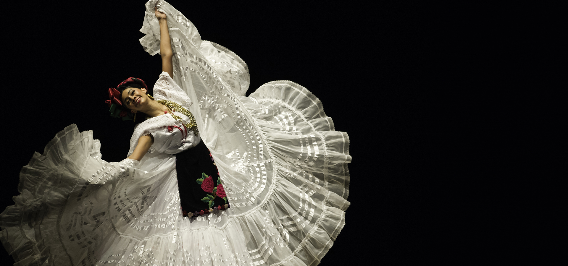 A solo woman poses in a traditional Mexican ornate flowing white dress, with her arms spead wide and the skirt in her hands, and smiles into the camera.