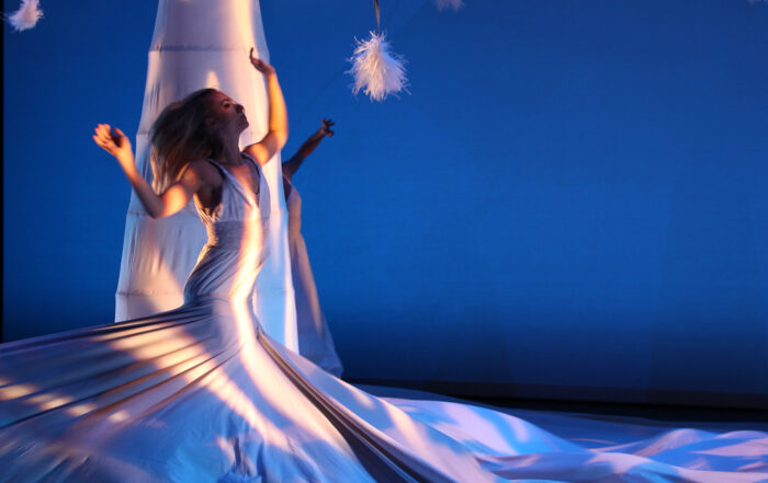 A solo female MOMIX dancer on stage in a wrapping, flowing white dress that covers most of the ground with her arms in the air and her head thrown back.