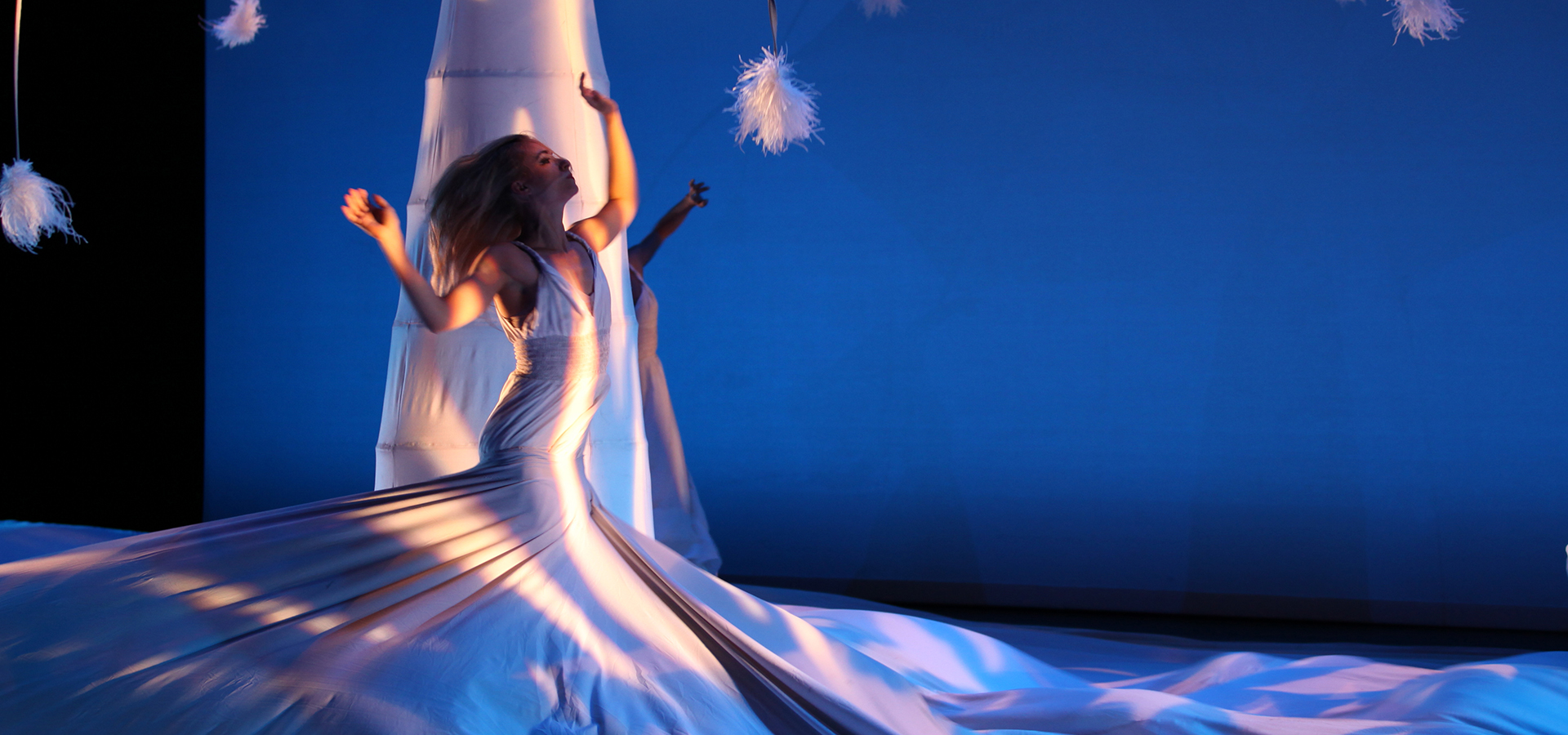 A solo female MOMIX dancer on stage in a wrapping, flowing white dress that covers most of the ground with her arms in the air and her head thrown back.