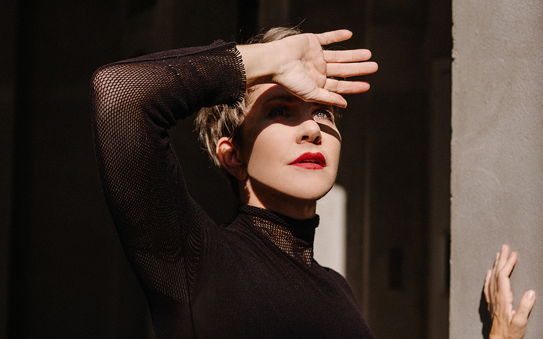 Mezzo-soprano Joyce DiDonato, a white woman with short light brown hair and wearing a black long-sleeve top, looks into the sun with an arm raised to shied her eyes.