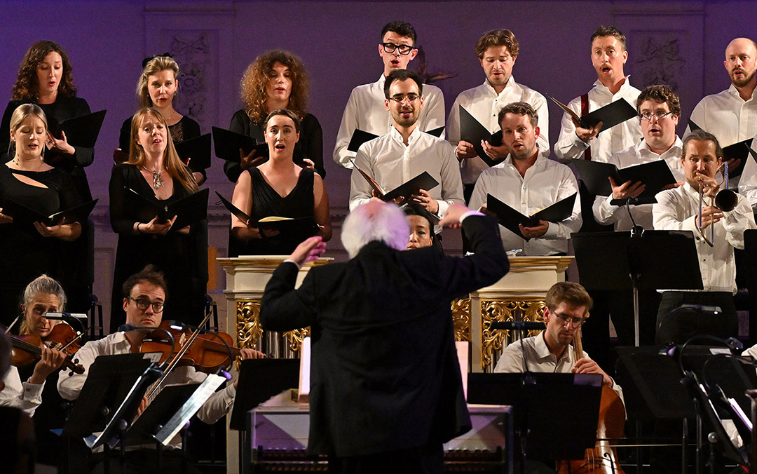 Conductor Masaaki Suzuki leads the Monteverdi Choir, a group of men and women, as they perform for an audience in a concert setting.