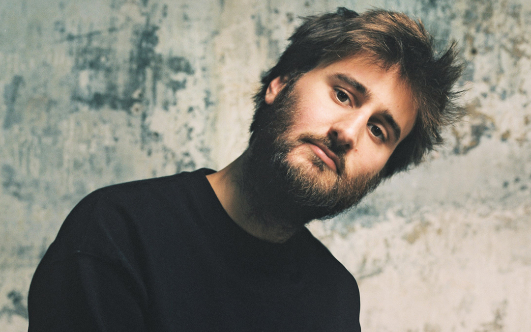Harpsichordist Jean Rondeau, a white man with brown hair and a beard, wearing a black crew-neck shirt, leaning and tilting his head to the right, looks into the camera against a mottled background.