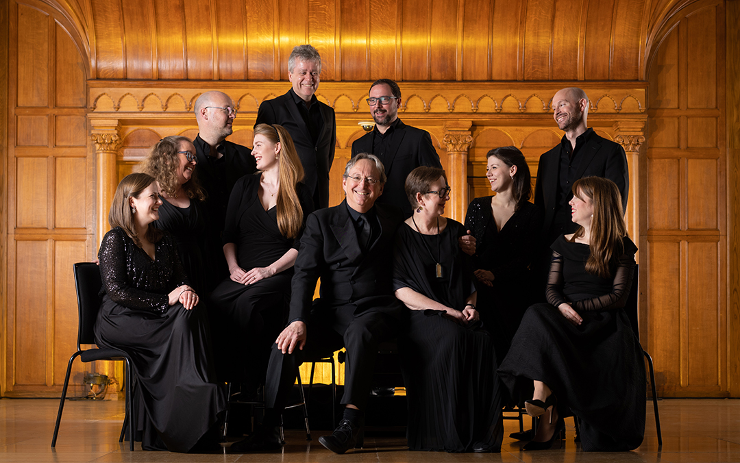 An enseble photo of the Tallis Scholars, a group of white men and women wearing black, all smiling and looking at each other against a golden wood background.