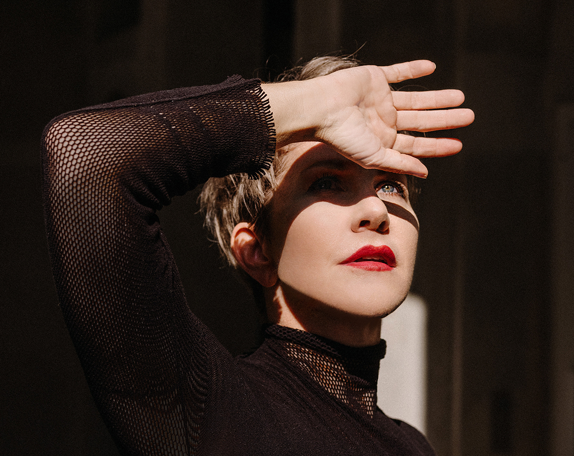 Mezzo-soprano Joyce DiDonato, a white woman with short light brown hair and wearing a black long-sleeve top, looks into the sun with an arm raised to shied her eyes.
