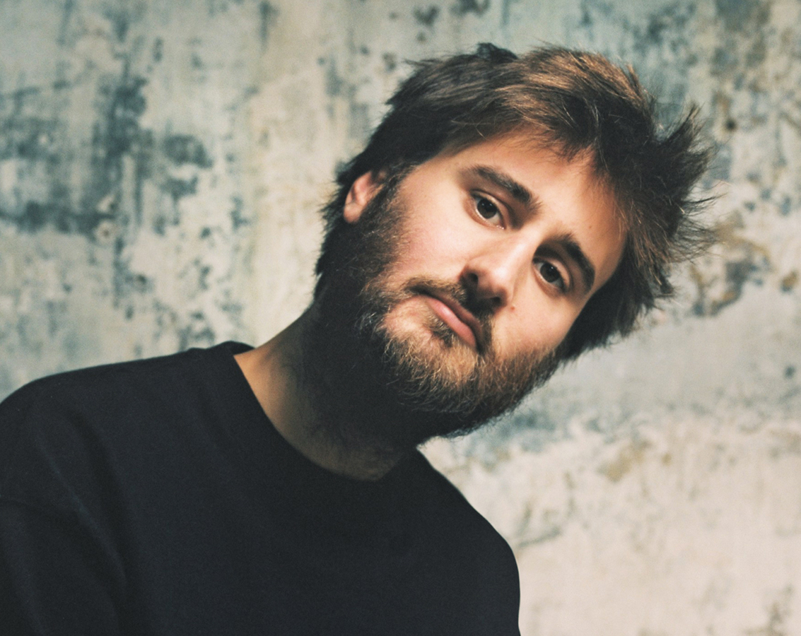 Harpsichordist Jean Rondeau, a white man with brown hair and a beard, wearing a black crew-neck shirt, leaning and tilting his head to the right, looks into the camera against a mottled background.