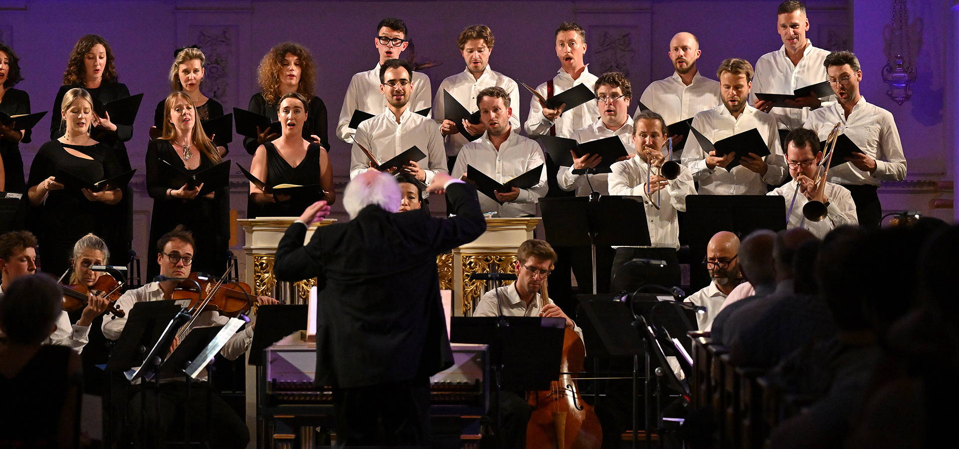 Conductor Masaaki Suzuki leads the Monteverdi Choir, a group of men and women, as they perform for an audience in a concert setting.