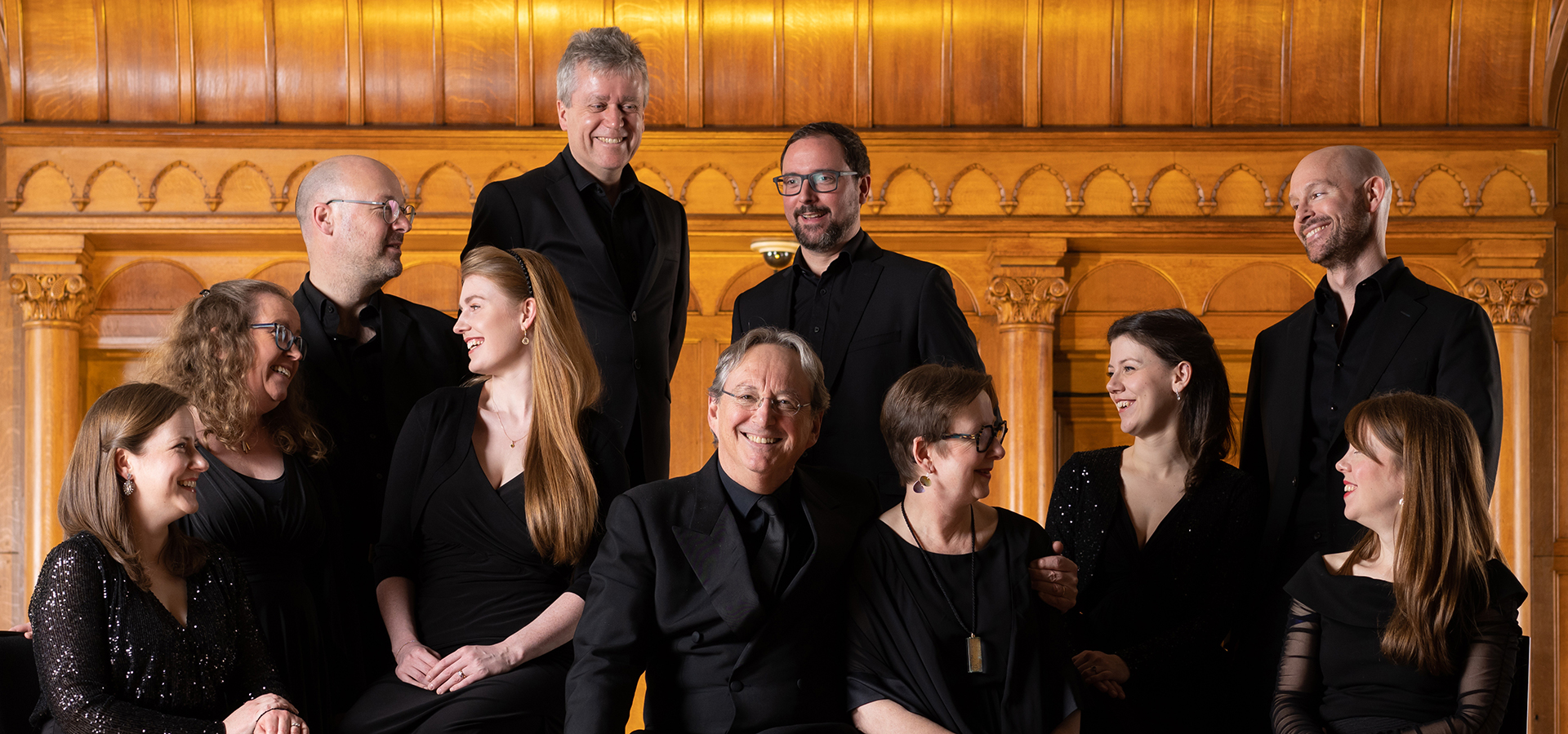 An enseble photo of the Tallis Scholars, a group of white men and women wearing black, all smiling and looking at each other against a golden wood background.
