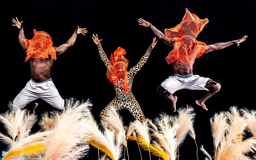 Three members -- two men and one woman -- of Cirque Kalabanté with bright orange transparent cloths over their heads and leaping into the air.