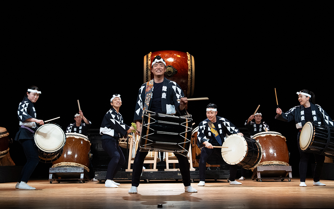A group of performers from Kodo playing multiple sized and shaped drums on stage.