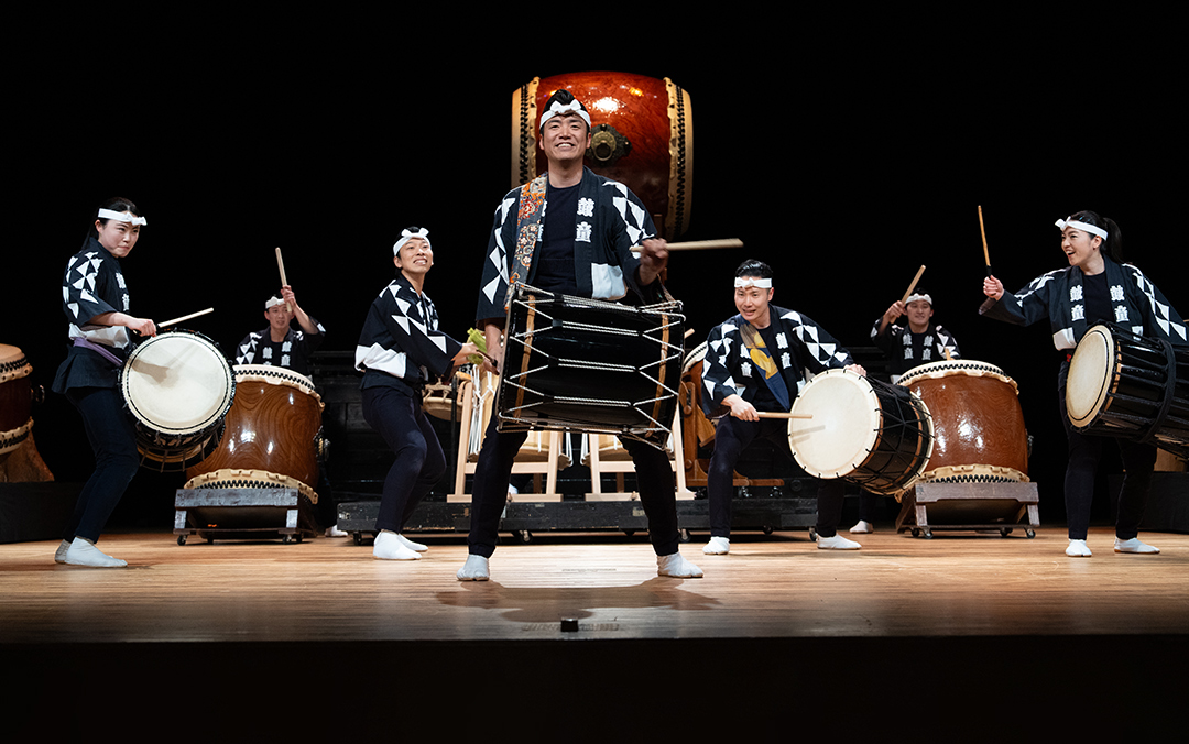 A group of performers from Kodo playing multiple sized and shaped drums on stage.