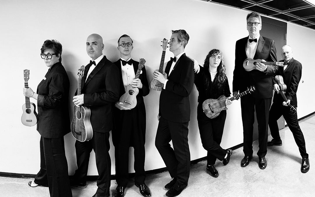 Black and white photo of members of Ukulele Orchestra of Great Britain all in line holding their instruments.