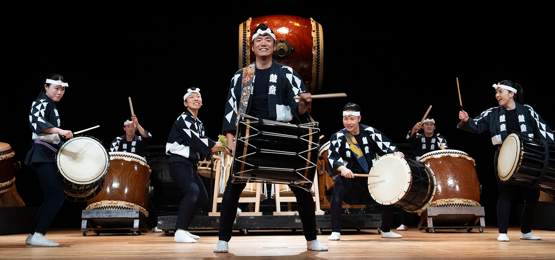 A group of performers from Kodo playing multiple sized and shaped drums on stage.