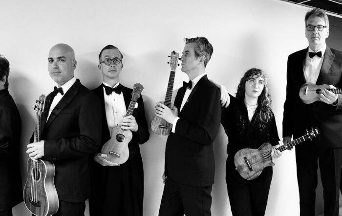 Black and white photo of members of Ukulele Orchestra of Great Britain all in line holding their instruments.