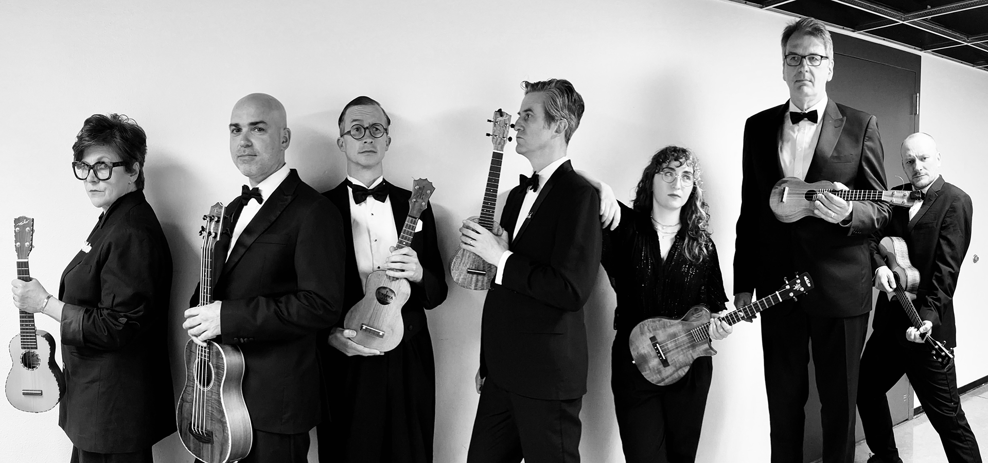 Black and white photo of members of Ukulele Orchestra of Great Britain all in line holding their instruments.