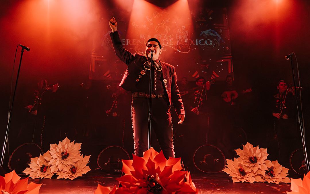A man dressed in a mariachi outfit performs on stage, adorned with flowers and bathed in red light, celebrating the culture of Mariachi Herencia de México.