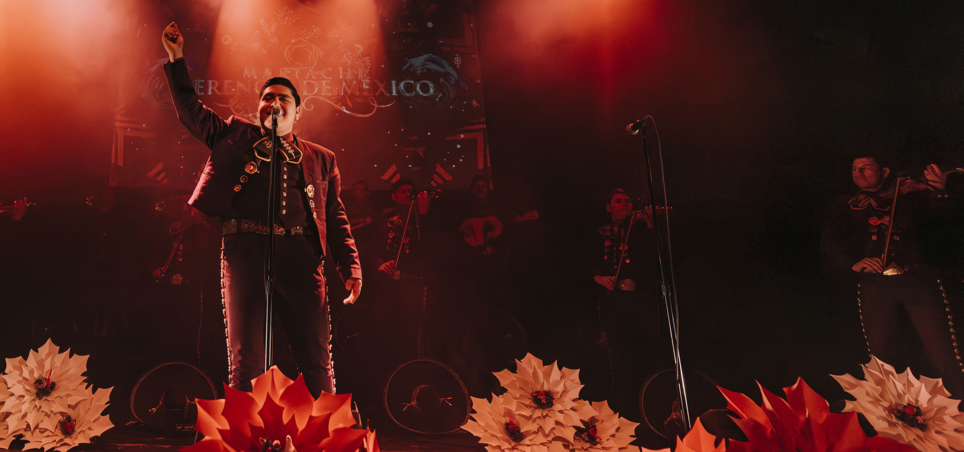 A man dressed in a mariachi outfit performs on stage, adorned with flowers and bathed in red light, celebrating the culture of Mariachi Herencia de México.