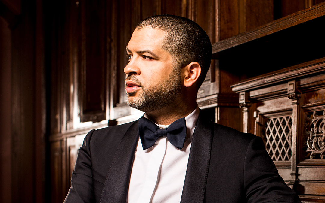Jazz pianist Jason Moran, a black man in a black suit and bow tie, looks to the side and leans agaist an upright piano.