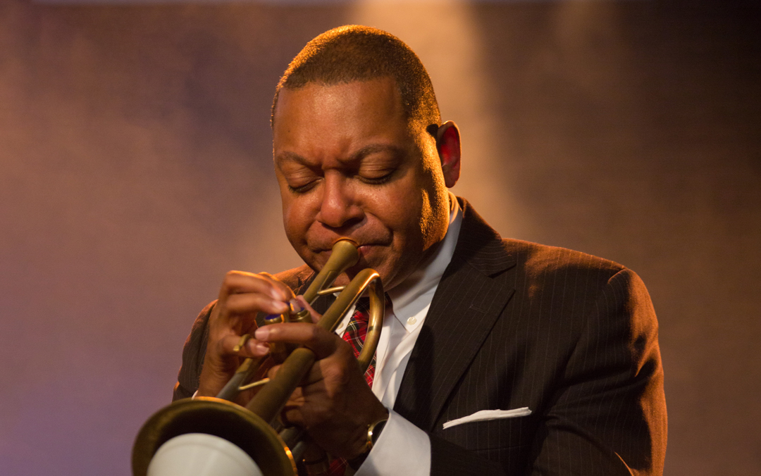 Famed trumpeter Wynton Marsalis of Jazz at Lincoln Center Orchestra, a black man in a dark suit and red tie playing his instrument.