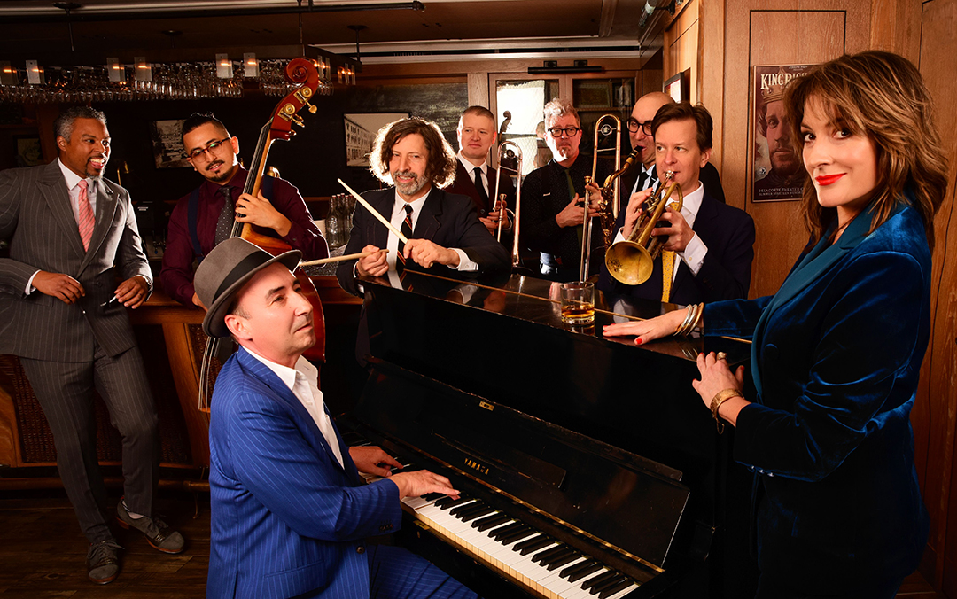 An ensemble photo of the Hot Sardines, a jazz group of eight men and one woman gathered around a piano with their instruments.